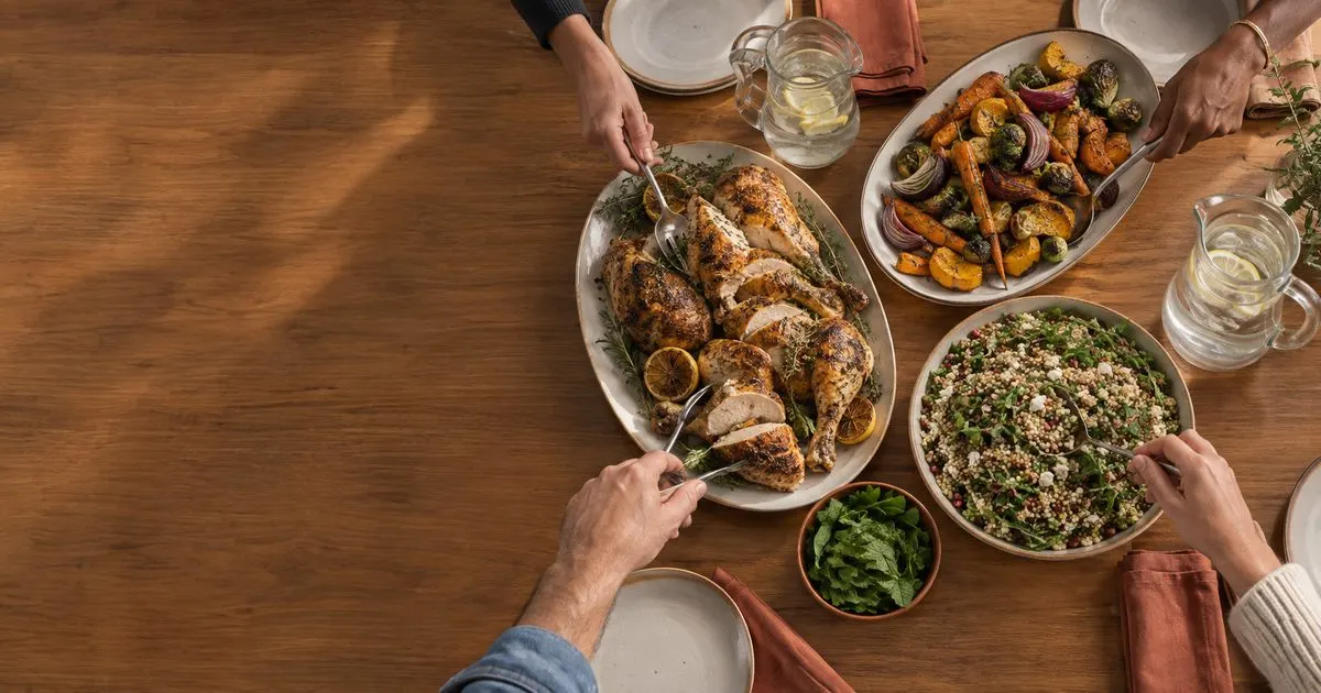 Hands reach toward platters of roast chicken, vegetables and salad on the right of a wooden table, leaving the left side empty.
